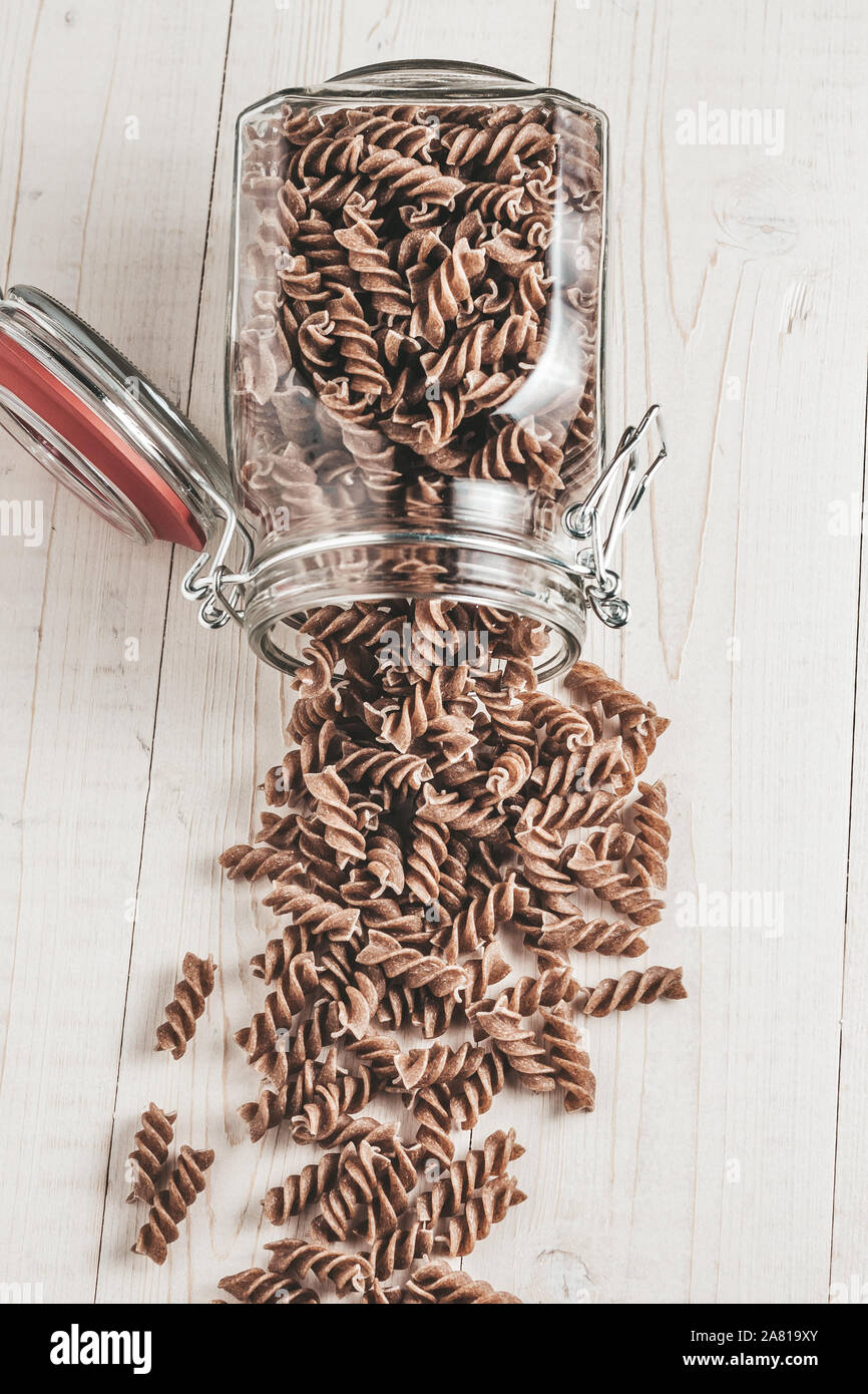 Maccheroni fusilli versata da un aperto il vasetto di vetro su un bianco tavolo in legno Foto Stock