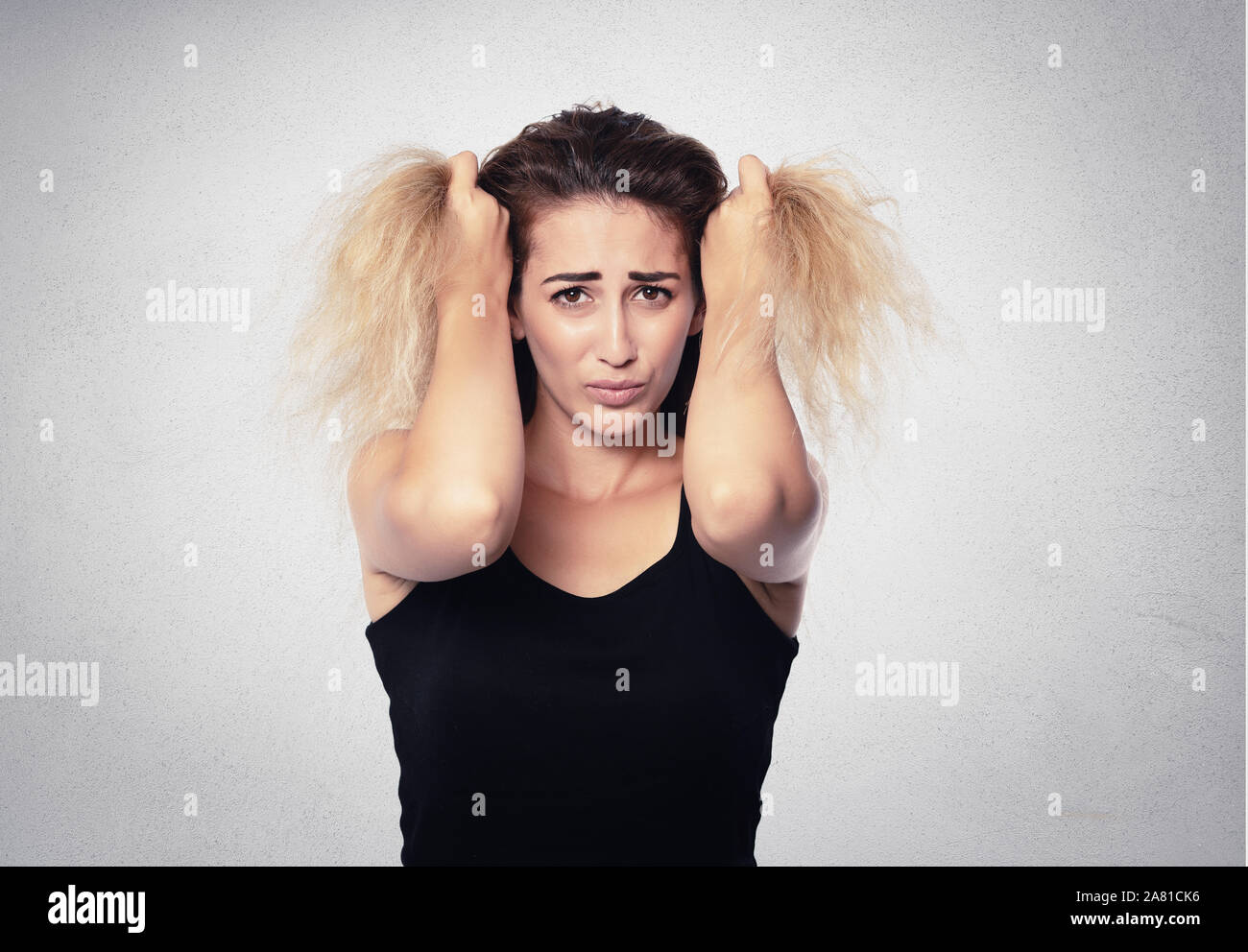 Sconvolto donna bionda tenendo i capelli sul muro grigio Sfondo . Foto Stock