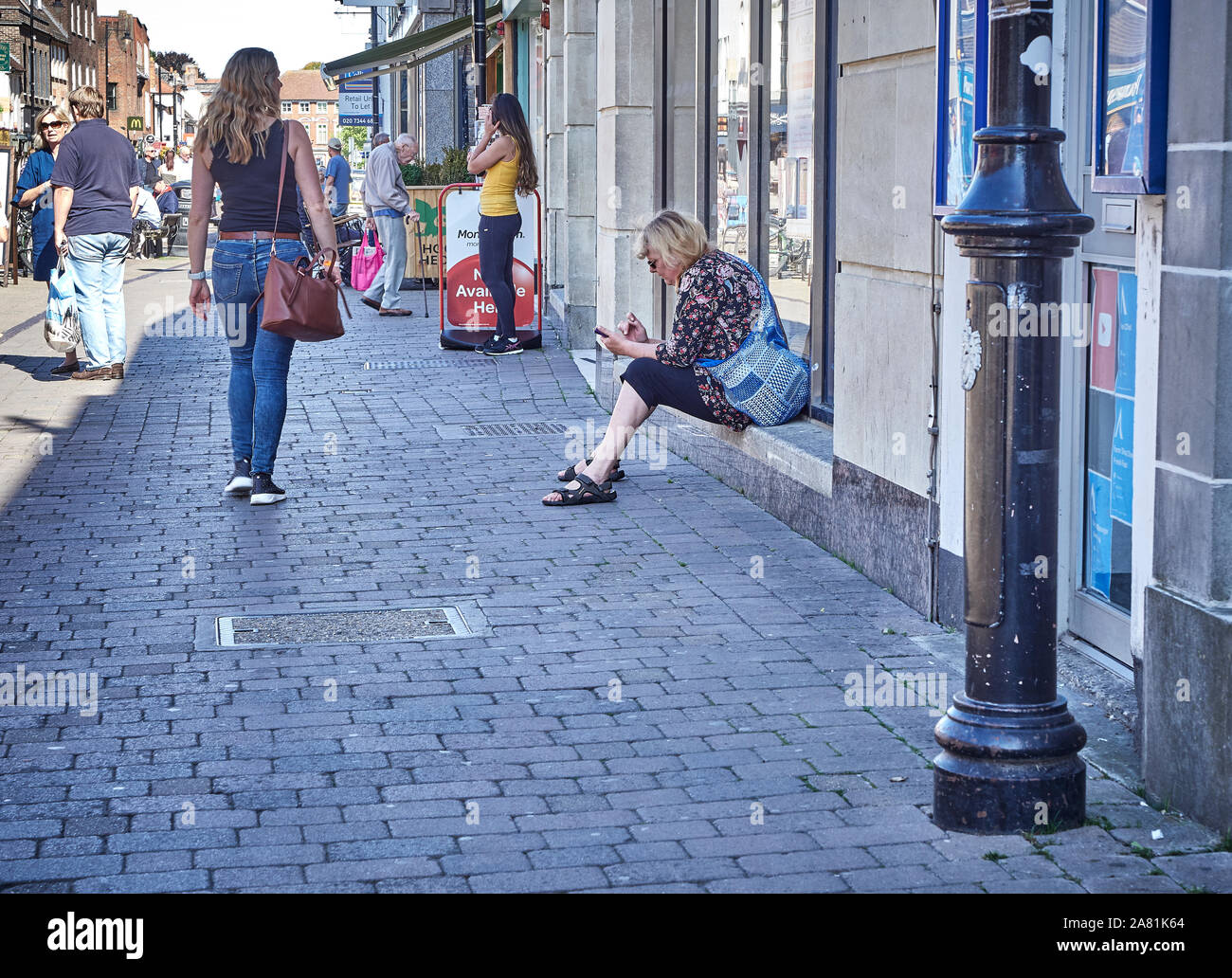 Una donna si sedette sul cill alla vetrina di un negozio utilizzando il proprio telefono cellulare come un altro donne passeggiate in passato Northbrook Street, Newbury, Regno Unito Foto Stock