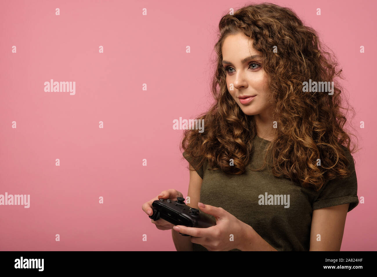 Piuttosto gamer ragazza con i capelli ricci azienda controller di gioco e concentrati durante la riproduzione di un videogioco isolato su sfondo rosa in studio Foto Stock