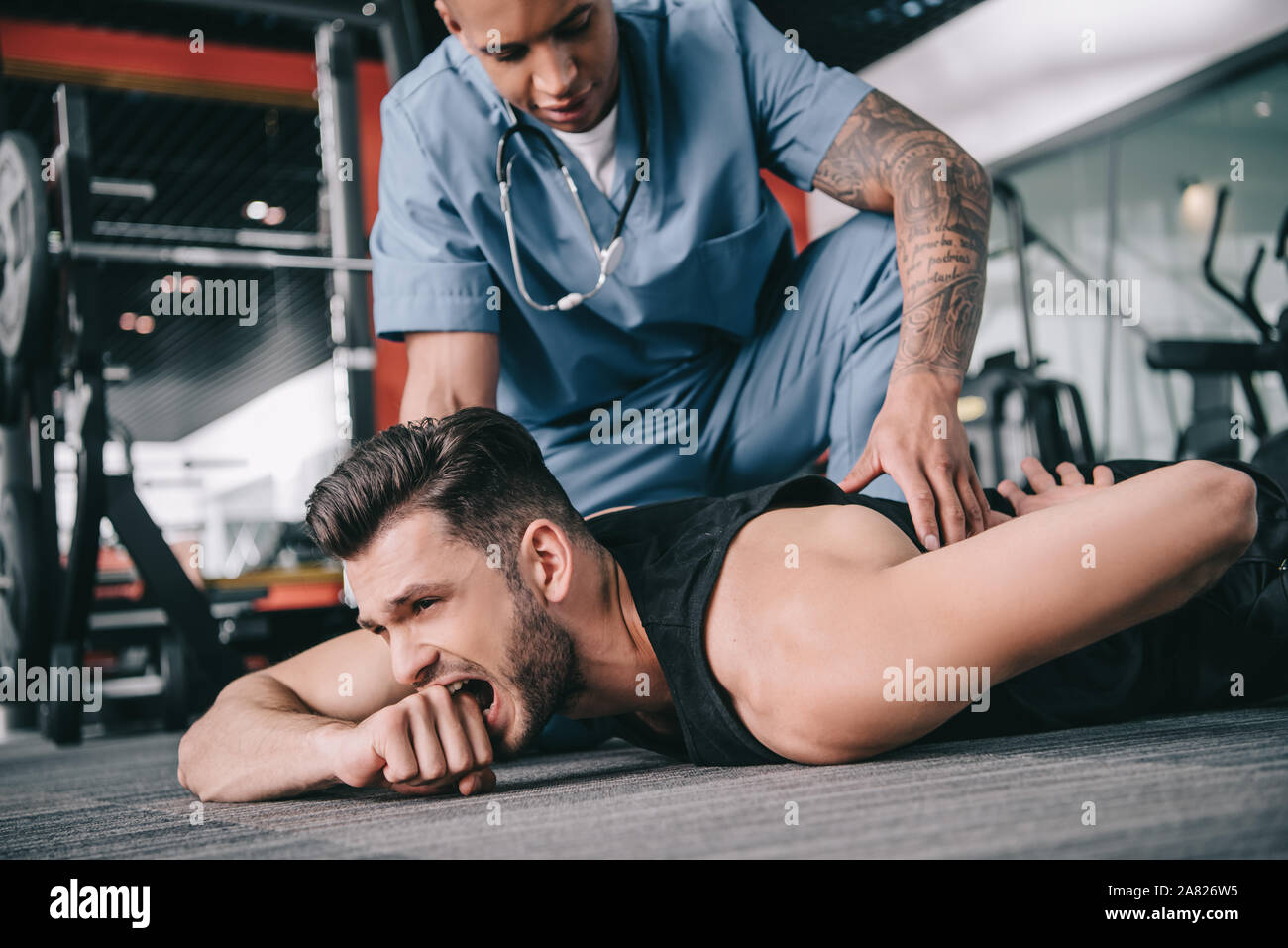 Giovane medico americano toccando feriti indietro di uno sportivo sdraiato sul pavimento e che soffrono di dolore Foto Stock