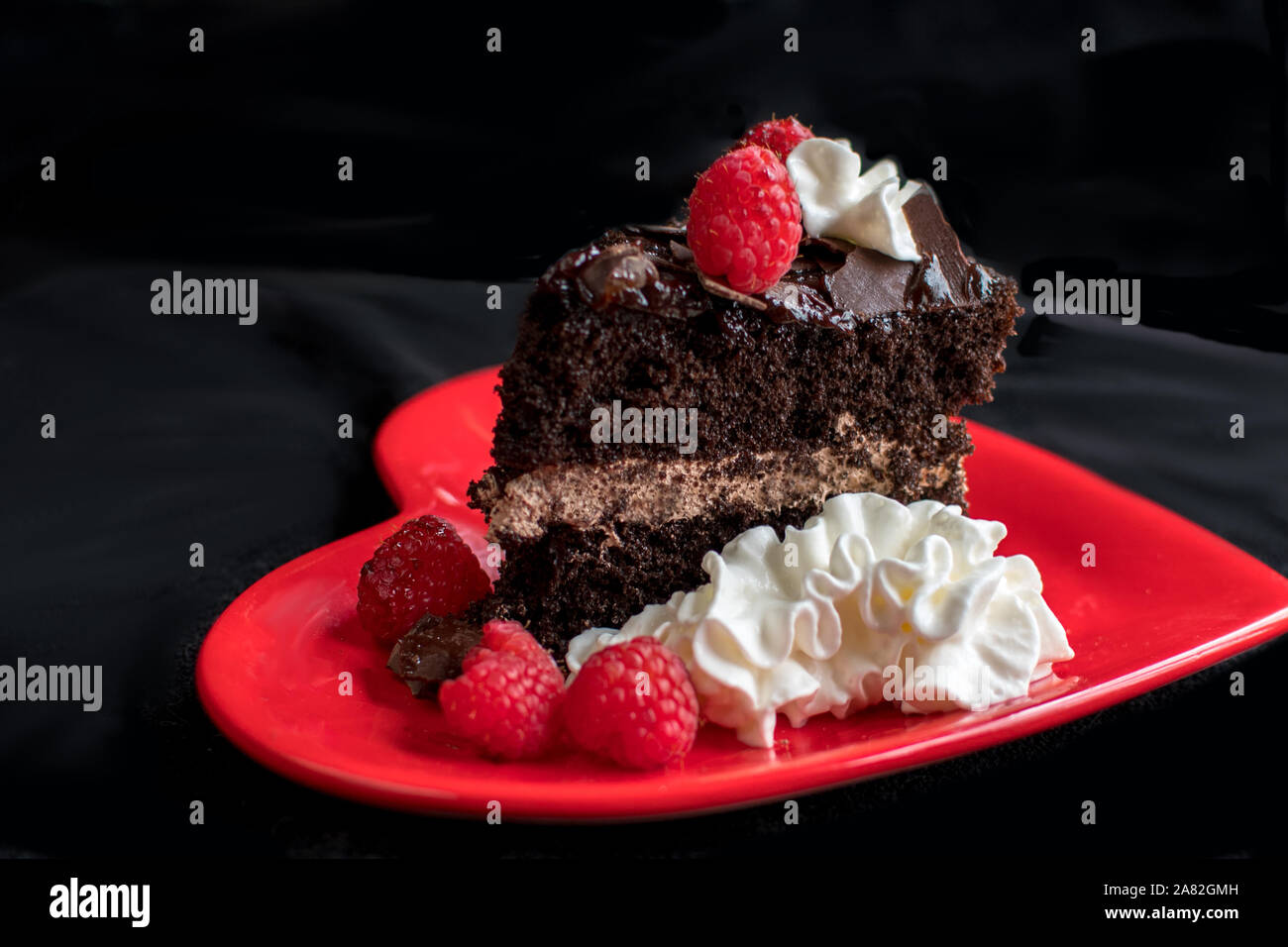 Una bella fetta di torta al cioccolato, poggia su un cuore rosso piastra, e guarnita con panna montata, lamponi e scaglie di cioccolato Foto Stock