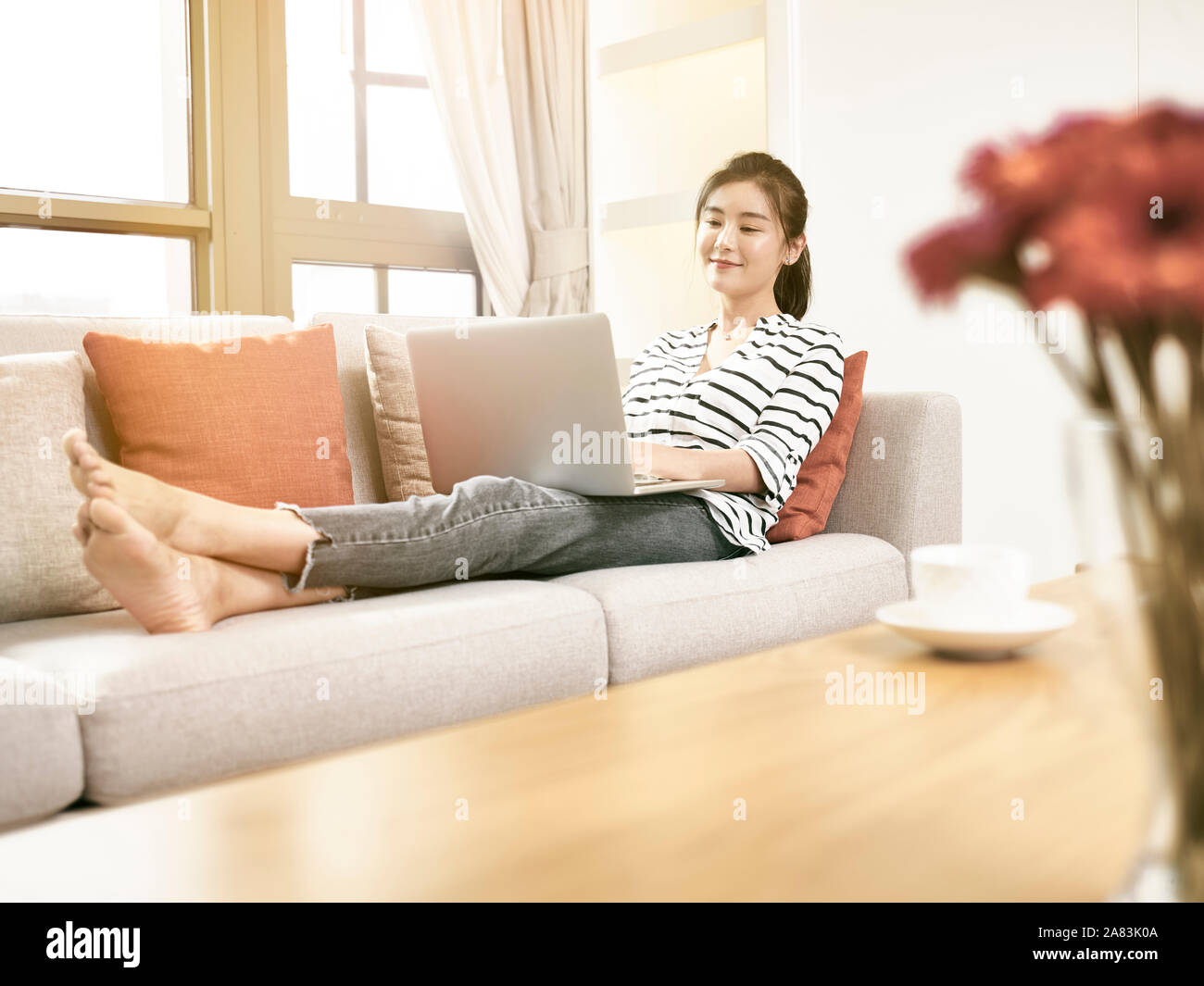 Bella e felice giovane donna asiatica di lavoro a casa sittng sul lettino usando il computer portatile Foto Stock