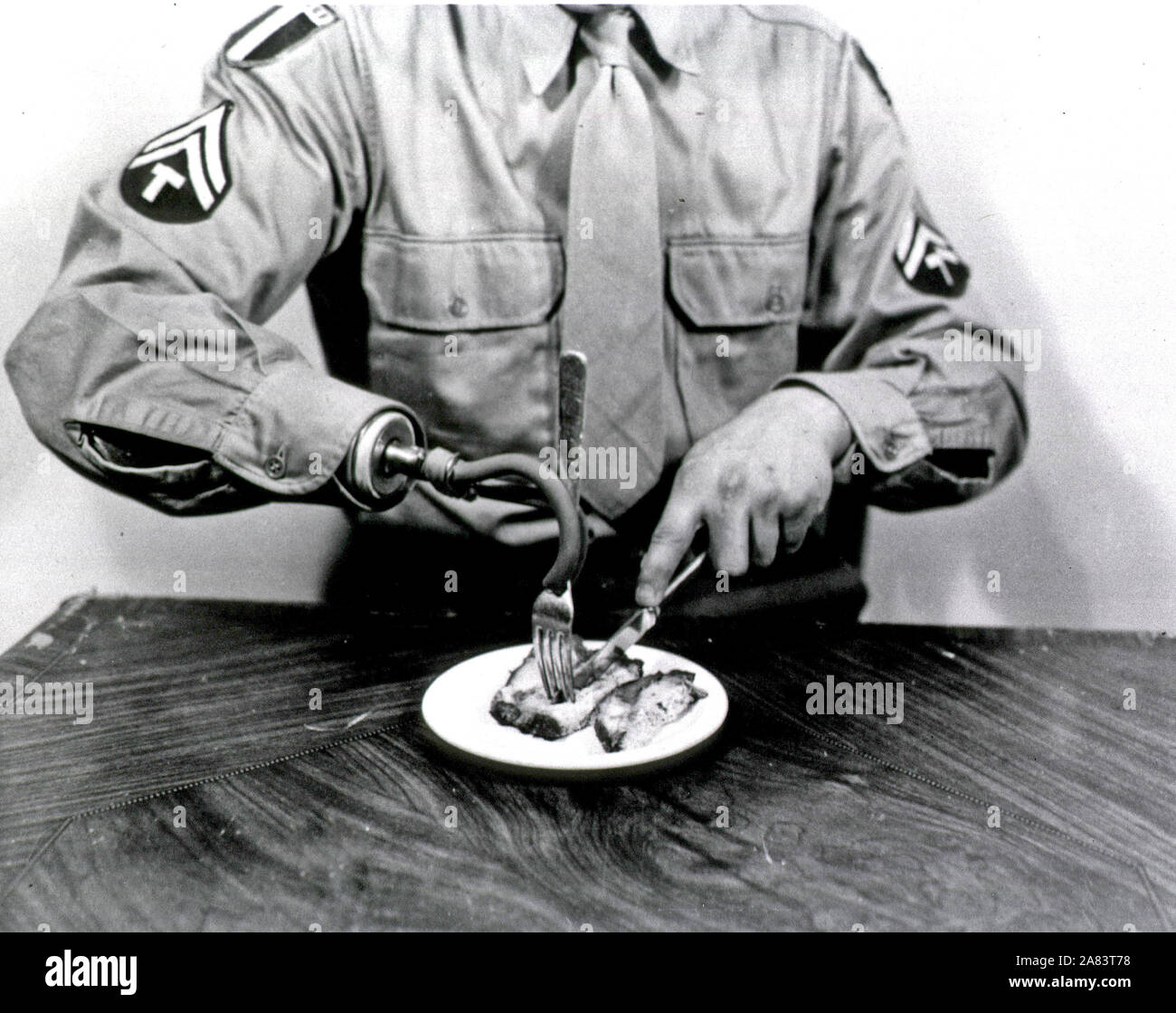 Vista di un uomo in uniforme militare rappresentata dal collo in giù seduti ad un tavolo. L'uomo detiene una forcella nella sua protesico gancio destro e con la sua mano sinistra taglia un pezzo di carne sul piatto. Foto Stock
