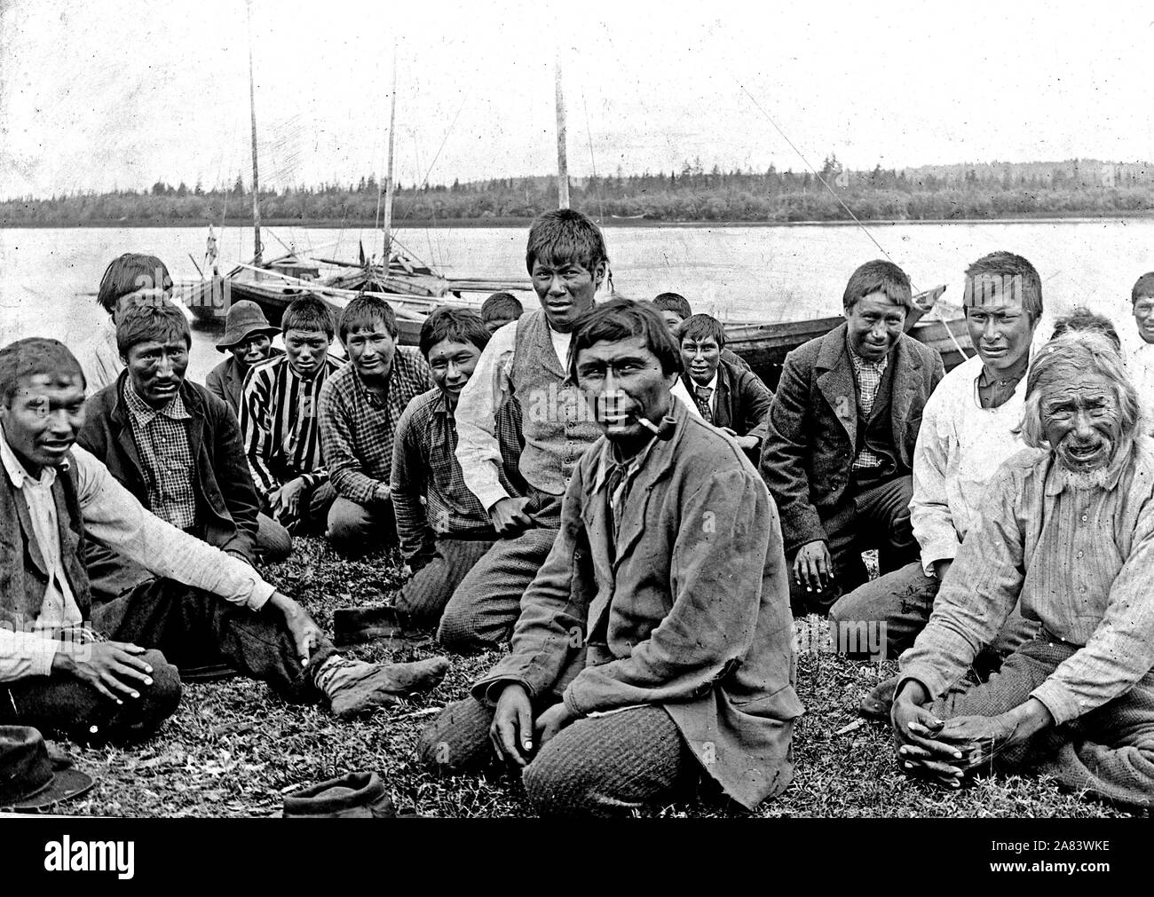 Indian uomini e ragazzi in abito occidentale, seduti lungo il litorale, barche in background. 1900-1923 della Baia di Hudson indiani Foto Stock