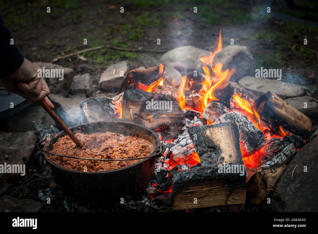 Una mano soffia una pentola di chili che cucinano sulle braci di un fuoco aperto. Foto Stock