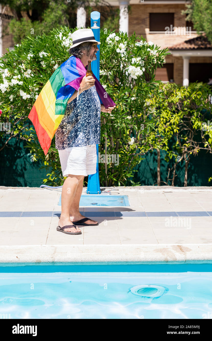 Stock Foto di un uomo con pantaloni corti e maglietta stampata, con una birra in mano e una bandiera arcobaleno accanto ad una piscina. Concetto Lgtb Foto Stock