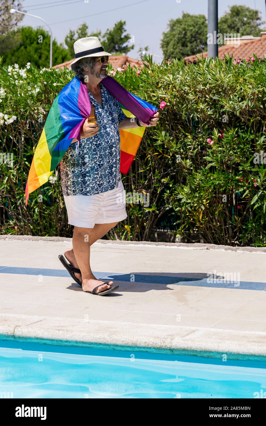 Stock Foto di un uomo con pantaloni corti e maglietta stampata a piedi con una birra in mano e una bandiera arcobaleno sul retro vicino a una piscina. Estate e tolleranza Foto Stock