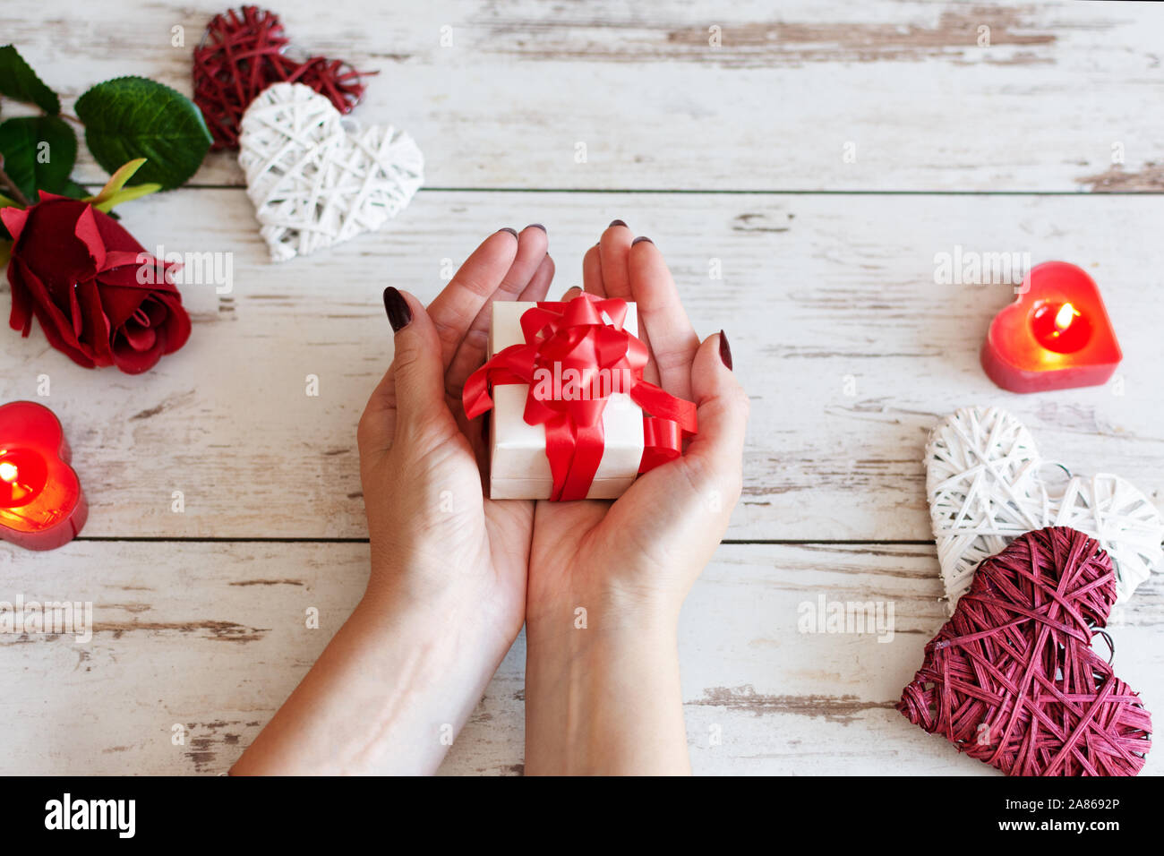 Regalo di San Valentino. La bellezza della donna mani confezione regalo con fiocco rosso su sfondo di vacanza con candele incandescente, fiori di rose e cuori. Foto Stock