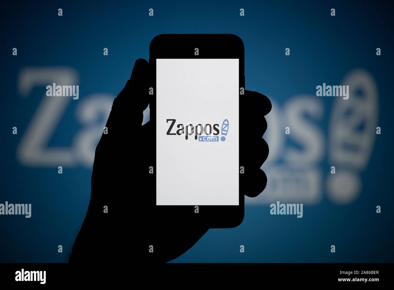 Un uomo guarda al suo iPhone che visualizza il logo Zappos, con lo stesso logo in background (solo uso editoriale). Foto Stock