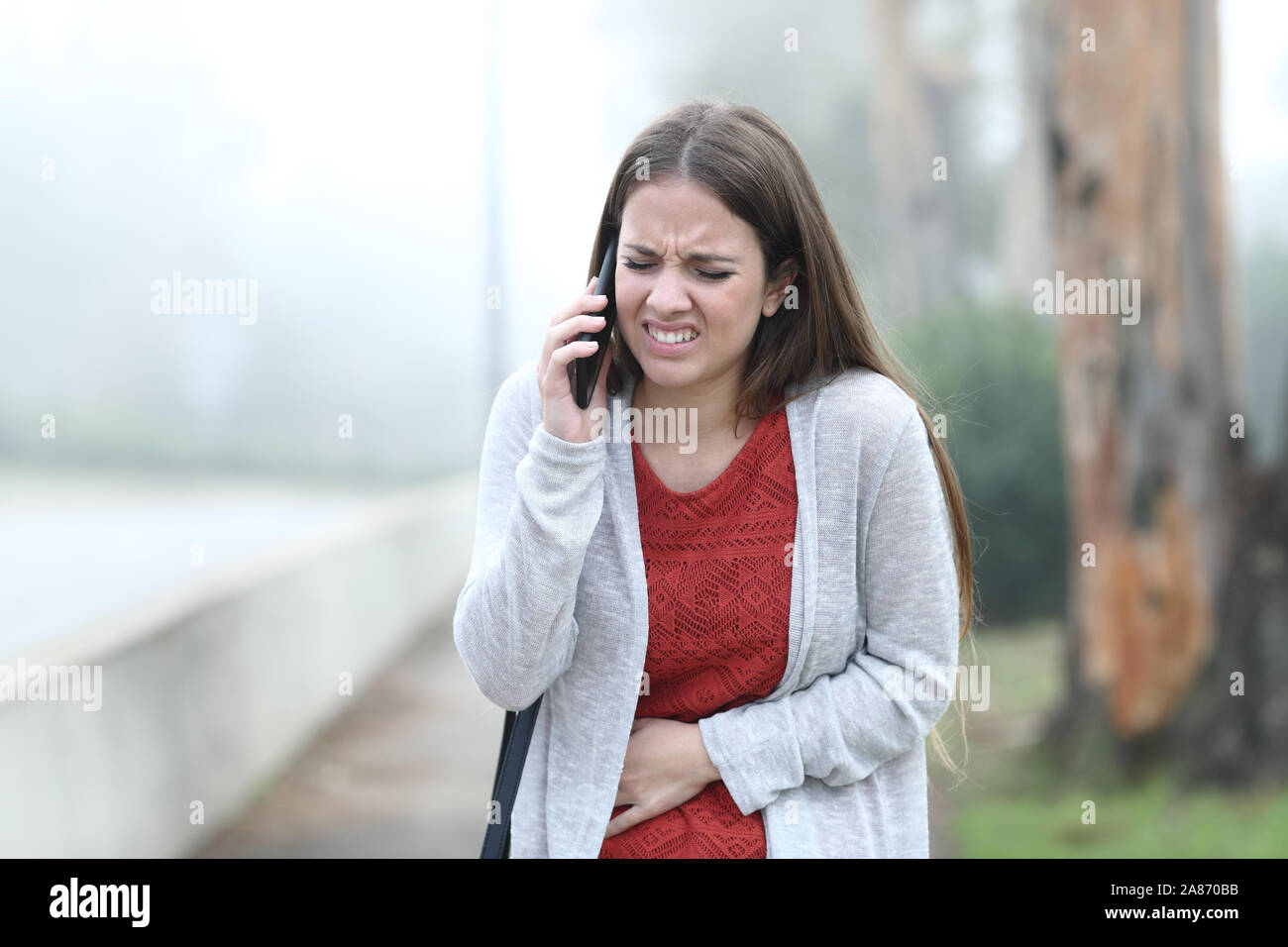 Vista frontale ritratto di una donna malata sofferenza mal di pancia chiamando al telefono in un parco Foto Stock