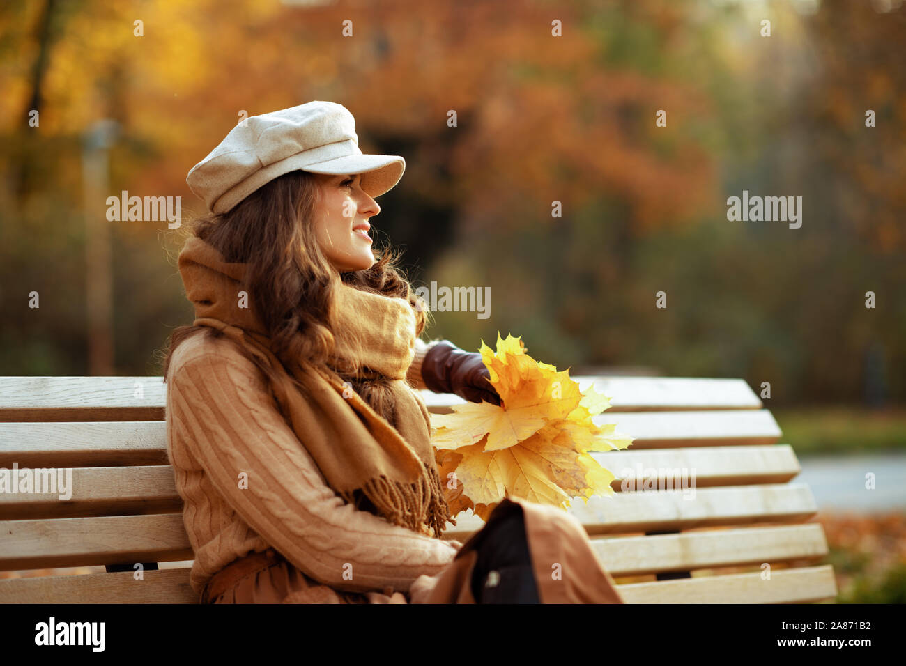 Ciao autunno. sorridente donna alla moda in un maglione, gonna, cappello, guanti e sciarpa con foglie di giallo guardando in lontananza mentre è seduto su una panchina outs Foto Stock