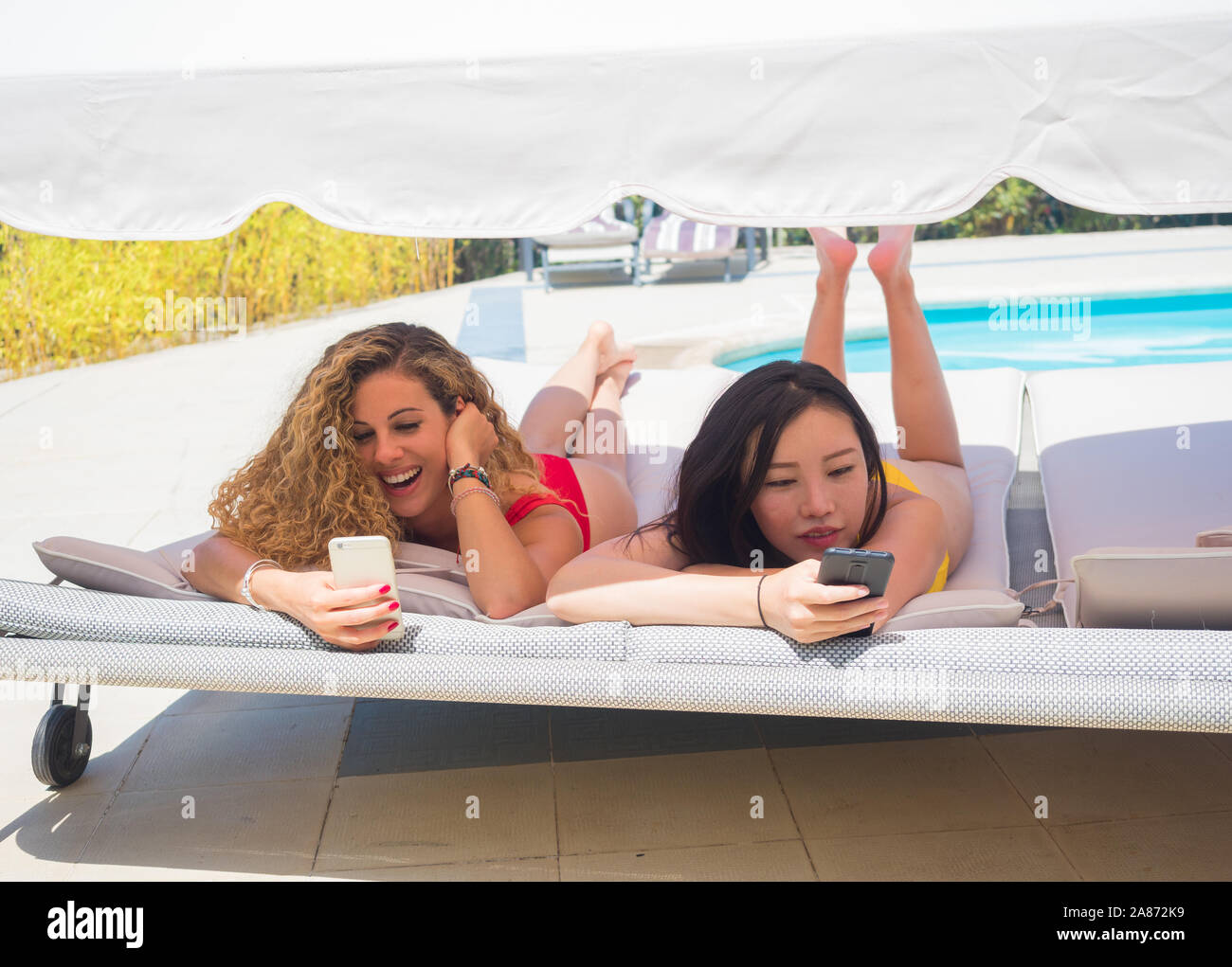 La donna caucasica e donna cinese giacente su una sedia a sdraio guardando i loro telefoni cellulari e ridere Foto Stock