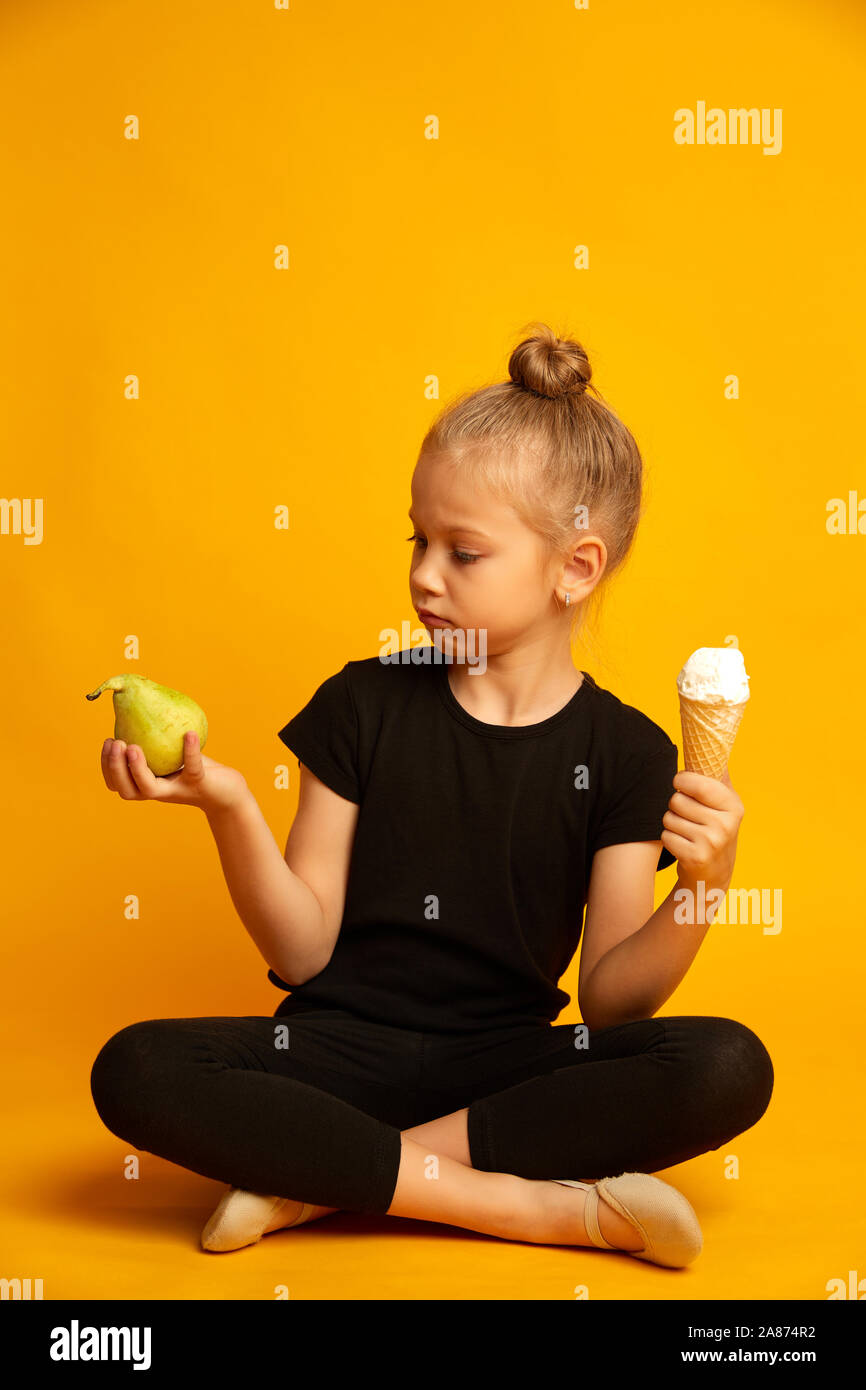 Incantevole piccola ragazza bionda scegliendo tra la pera e il dolce di gelati su sfondo giallo Foto Stock