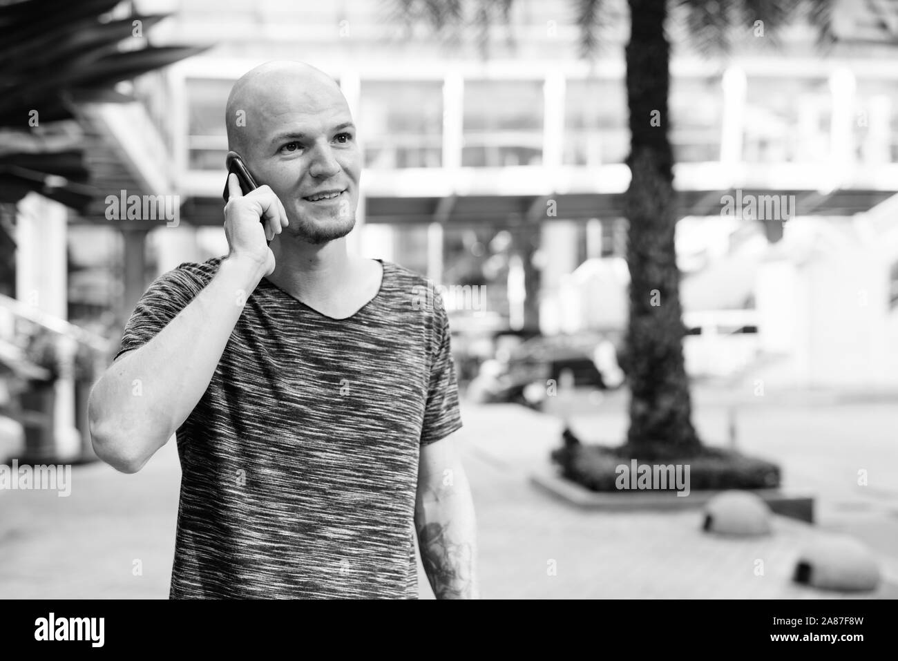 Felice giovane uomo calvo di pensare e parlare al cellulare al di fuori del centro commerciale Foto Stock