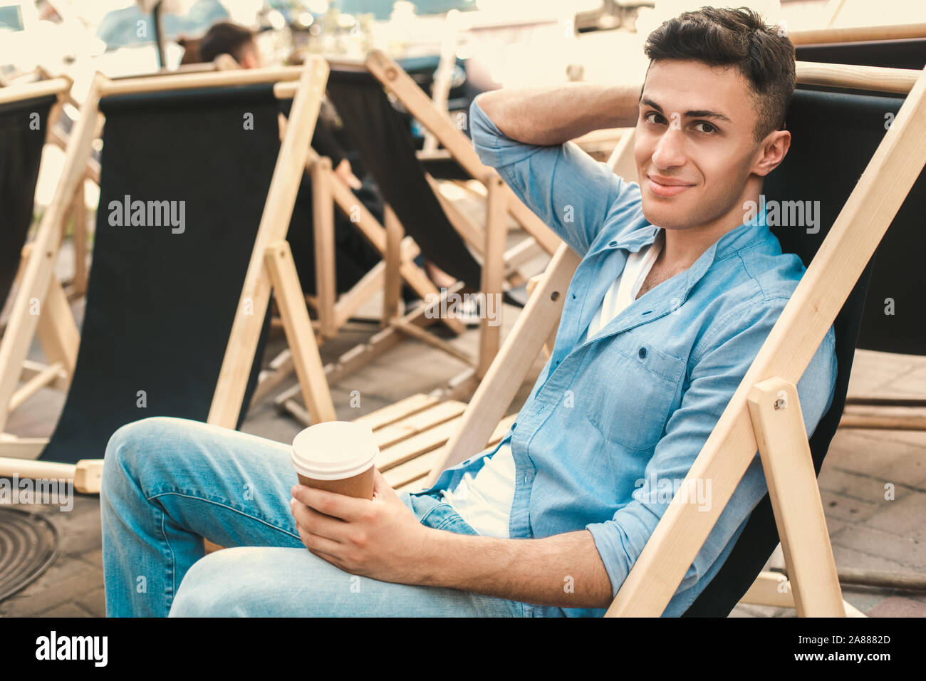 Per il tempo libero all'aria aperta. Elegante viaggiatore seduto sulla spiaggia con la tazza di caffè rilassato sorridente Foto Stock
