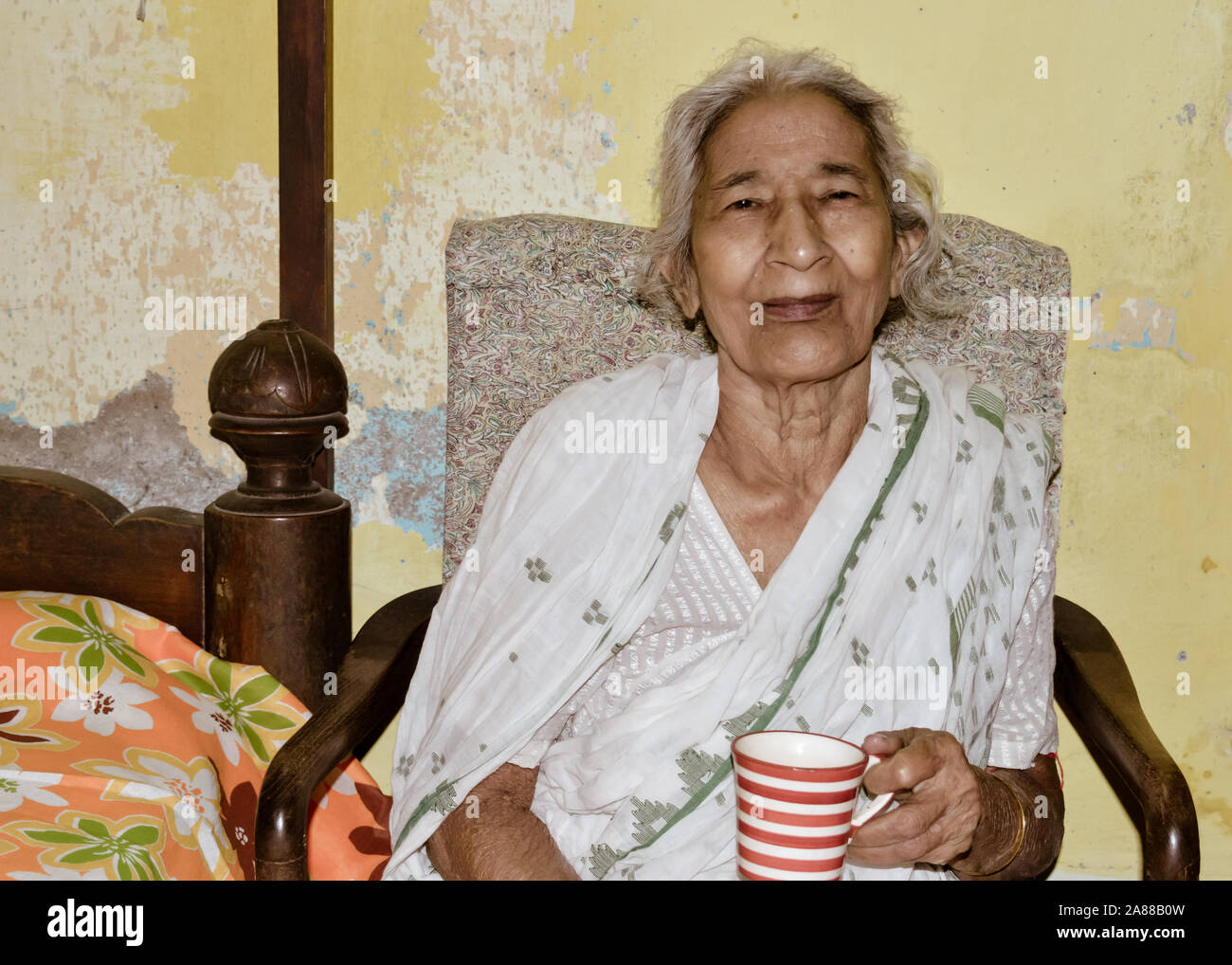 Ritratto di sorridere Active senior asiatici ed etnia indiana donna di 82 anni seduti su una sedia tenendo una tazza di caffè in appoggio a casa mentre cerca Foto Stock