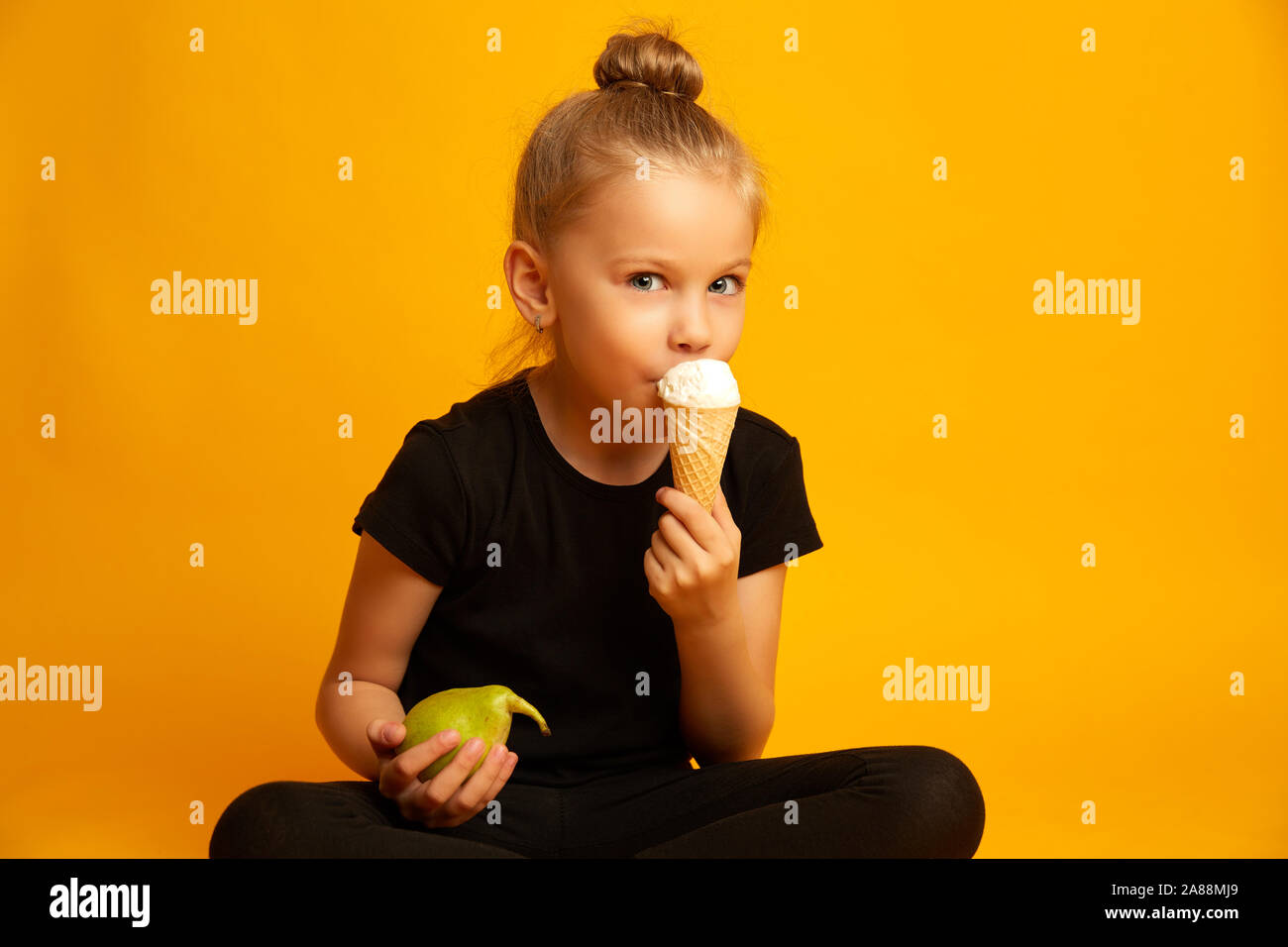 Disorientati bambina in body e scarpe da ballo scegliendo tra un sano e cibo malsano seduti gambe incrociate contro sfondo giallo Foto Stock
