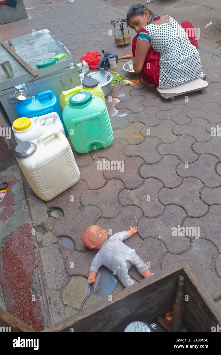 Una giovane donna il taglio di verdure su un marciapiede in Mumbai, India, guardando sopra la sua spalla a un bambini bambola giacente a terra Foto Stock