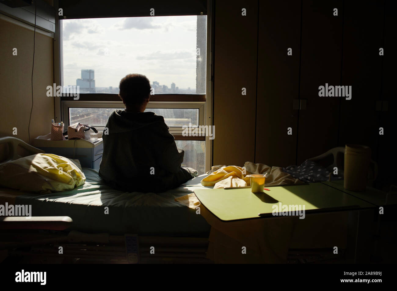 Donna seduta sul letto di ospedale, la schiena girata, guardando fuori della finestra - da soli Foto Stock