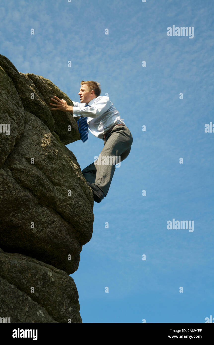 Valoroso uomo d affari in camicia e cravatta aggrappato al bordo di una pericolosa rocky Cliff Outdoors nel cielo blu Foto Stock