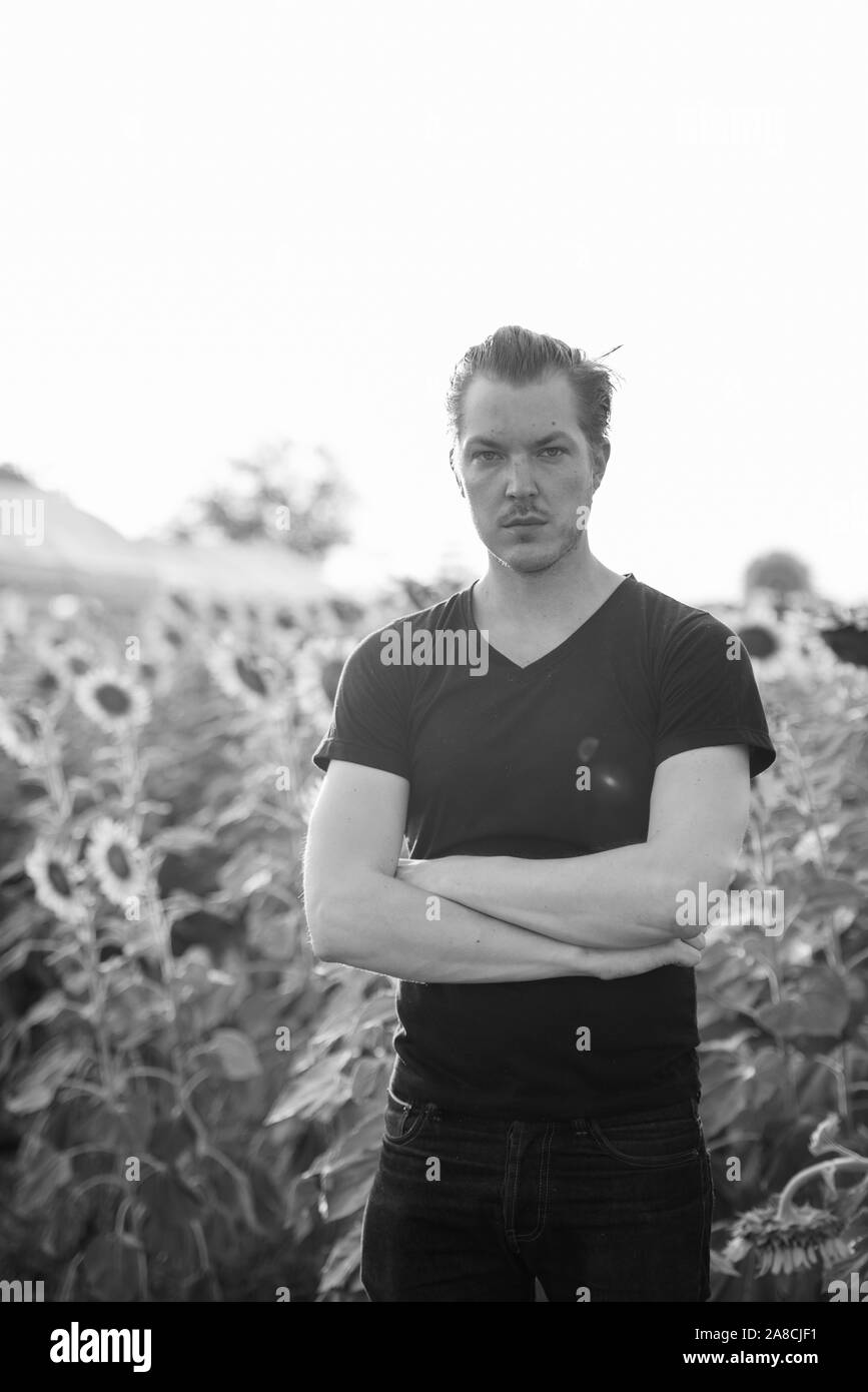 Giovane uomo bello in piedi con le braccia incrociate contro il campo di girasoli in fiore Foto Stock