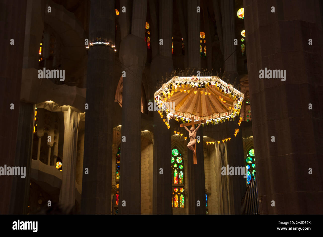 La Sagrada Familia - interno. Appendere il lampadario scultura di Gesù tra alti pilastri contro uno sfondo di finestre di vetro macchiate in colori caldi. Foto Stock