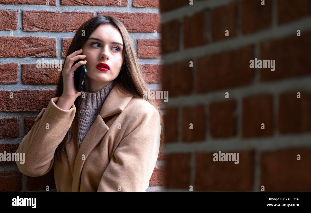 Ritratto di una ragazza sulla strada chiamando al telefono.una giovane donna siede contro un muro di mattoni e colloqui sul telefono. sul giovane ragazza autunno overc beige Foto Stock