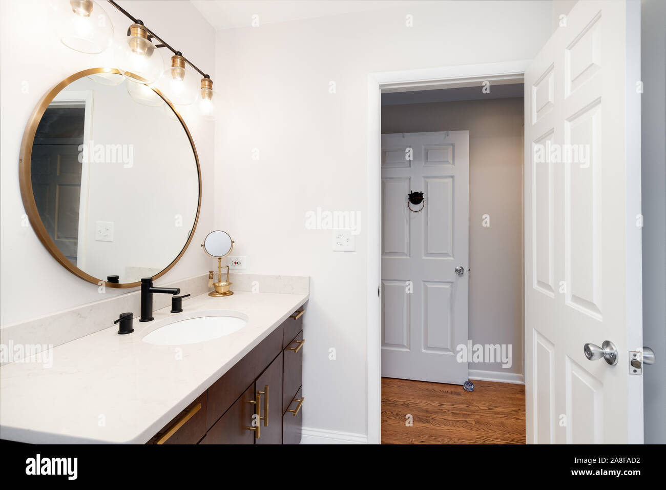 Un piccolo bagno con specchio circolare, moderno lampadario e ripiani in granito guardando verso il corridoio. Foto Stock