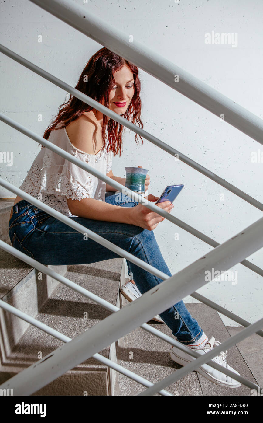 Giovane donna con i capelli rossi seduta sulle scale e parlando sul suo smartphone e di bere il caffè. Foto Stock