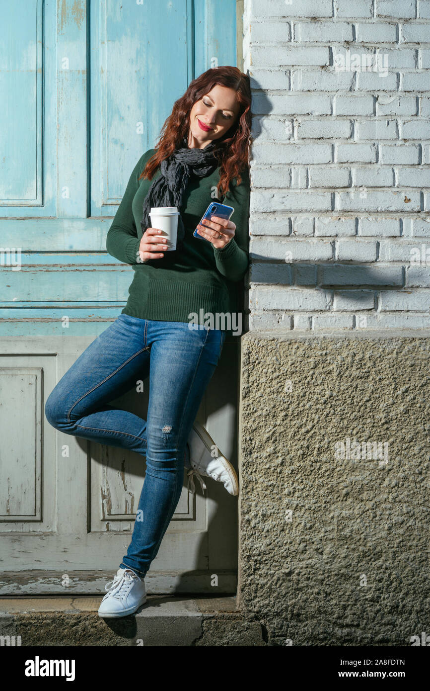Giovane donna con i capelli rossi guardando il suo cellulare e di bere il caffè da una tazza di asporto. Foto Stock