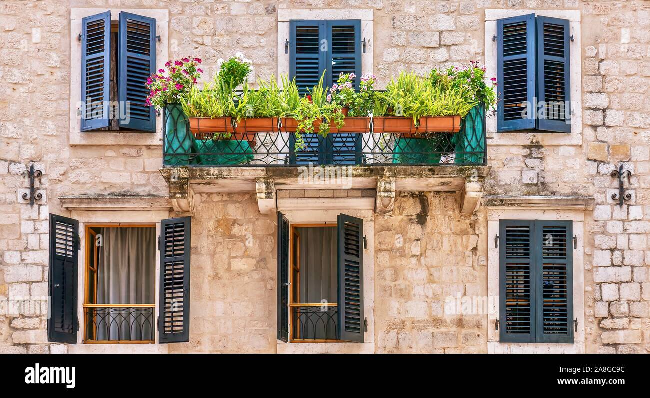 Vista sulla strada di una bella casa in pietra con facciata dipinta di persiane alle finestre e un piccolo balcone con una cancellata in ferro battuto e di piante in vaso. Montenegro. Foto Stock