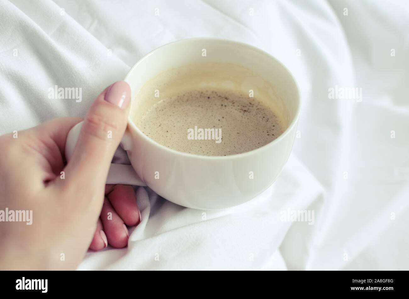 Una tazza di caffè in mano femminile nel letto su una coltre bianca. Moman godendo del comfort a casa accogliente atmosfera. Buona mattina. POV. Foto Stock