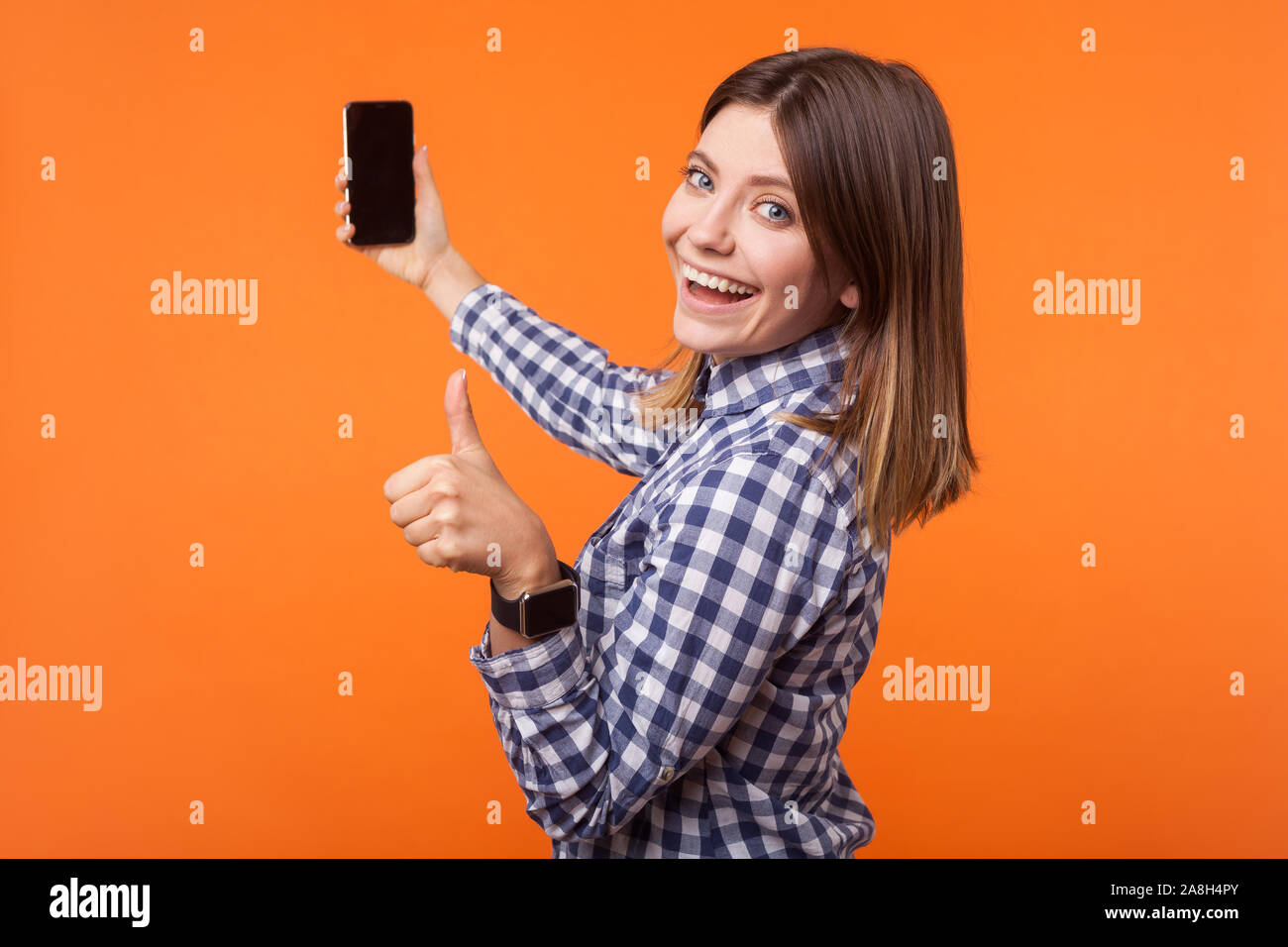Mezza girata ritratto di donna bruna con sorriso affascinante nella camicia  a scacchi tenendo il telefono e che mostra Thumbs up, soddisfatto con i  servizi di telefonia mobile. ind Foto stock -