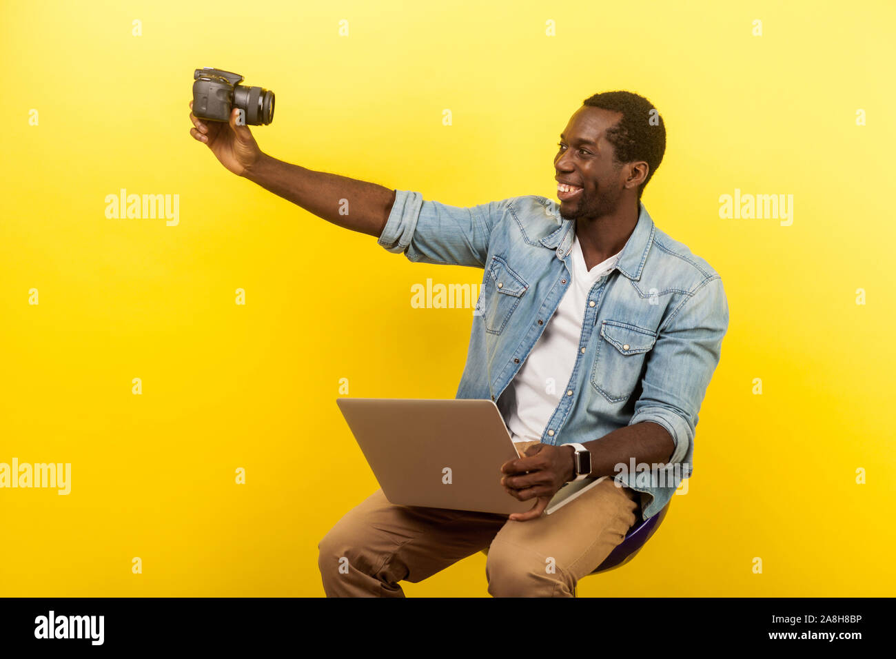 Ritratto di Allegro fotografo amatoriale con sorriso toothy in denim shirt tenendo selfie foto digitali con fotocamera DSLR e azienda portatile, di video live Foto Stock
