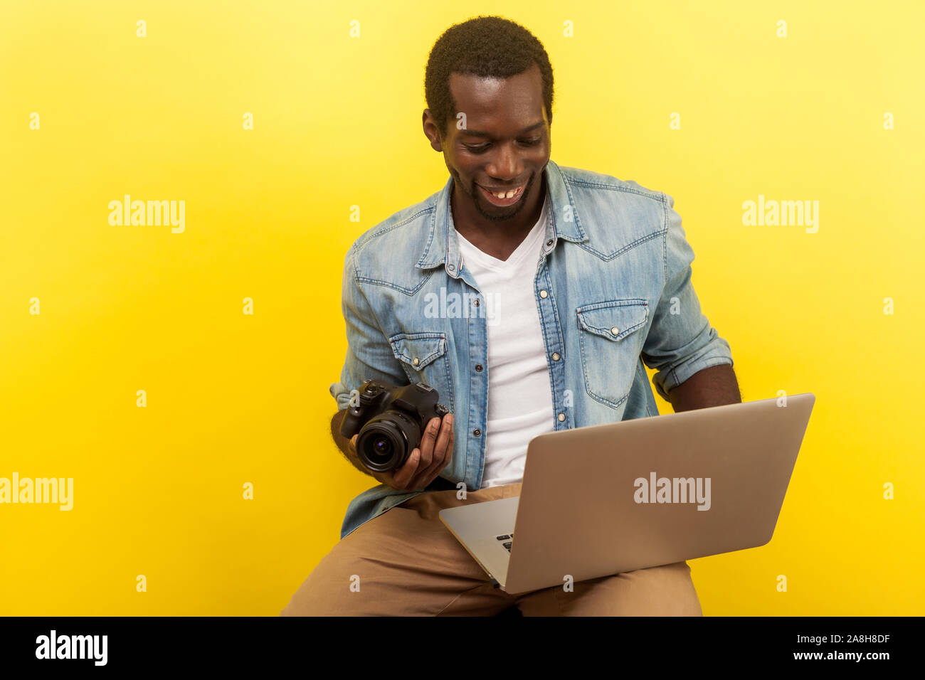 Ritratto del maschio positivo fotografo in denim shirt azienda digitale fotocamera DSLR e laptop, andando a prendere foto, utilizzando photo editing app su comput Foto Stock