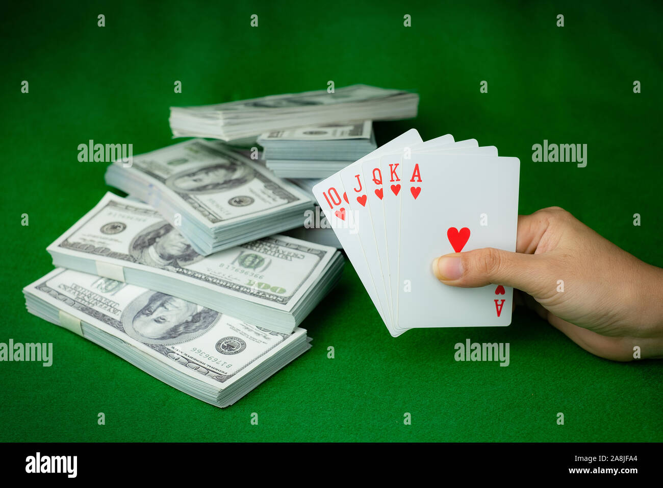 Poker Casino Gambling Royal Straight Flush e la pila di denaro di 100 dollari USA banconote presso lo sfondo verde, la mano che tiene la riproduzione di cuore di carta 5 Foto Stock