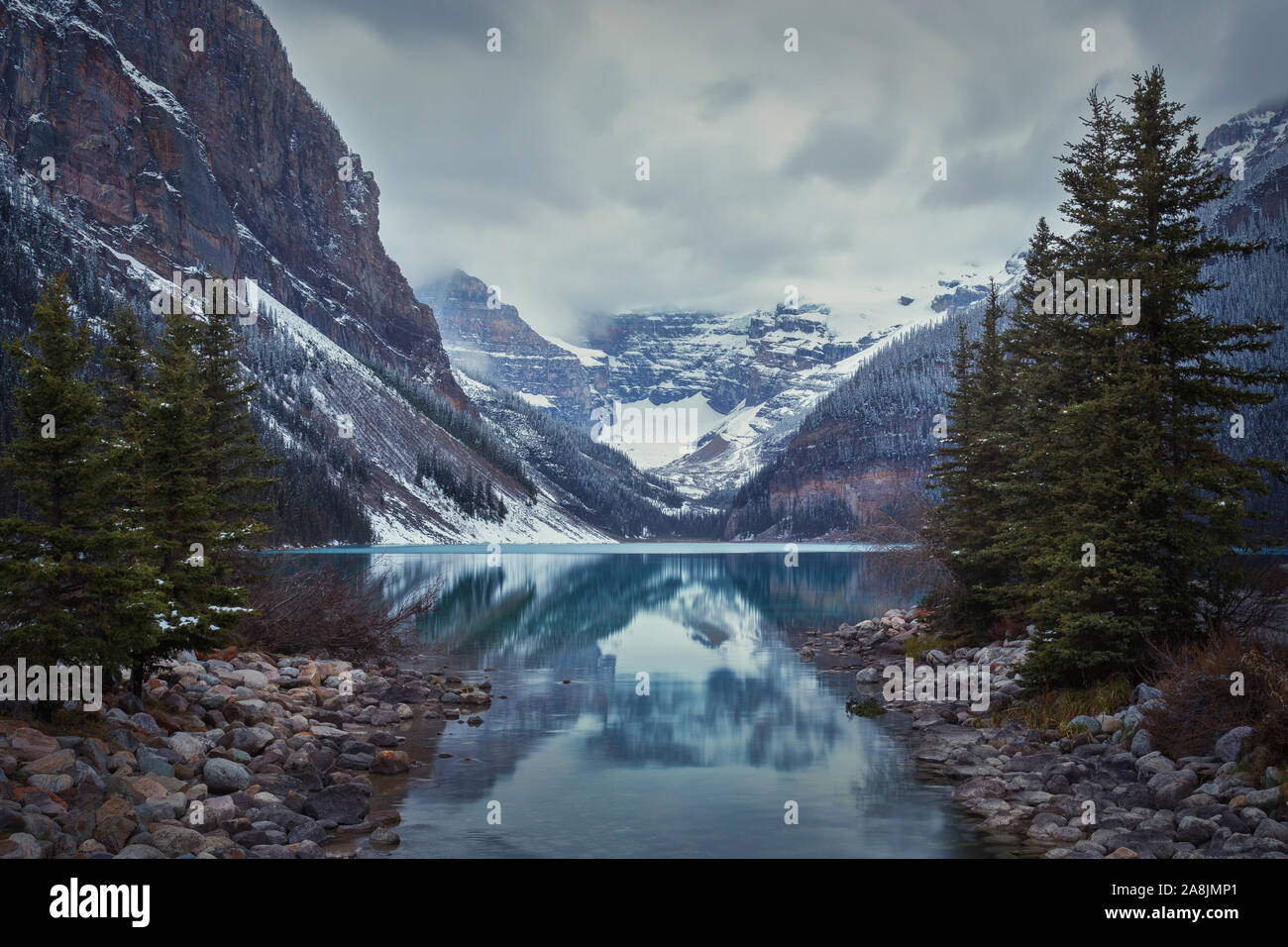 Il lago Louise in un giorno nuvoloso. La riflessione delle montagne innevate e pini sulle acque turchesi. Foto Stock