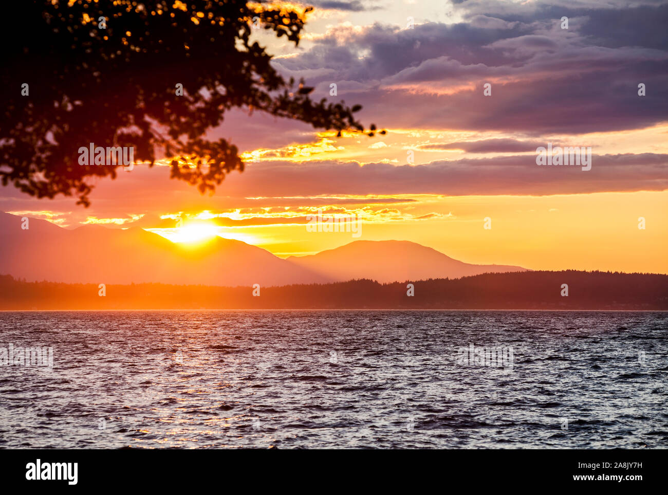 Il sole che tramonta oltre le montagne olimpiche e Puget Sound come visto da ovest vicino Seattle Alki Beach, Washington, Stati Uniti d'America. Foto Stock