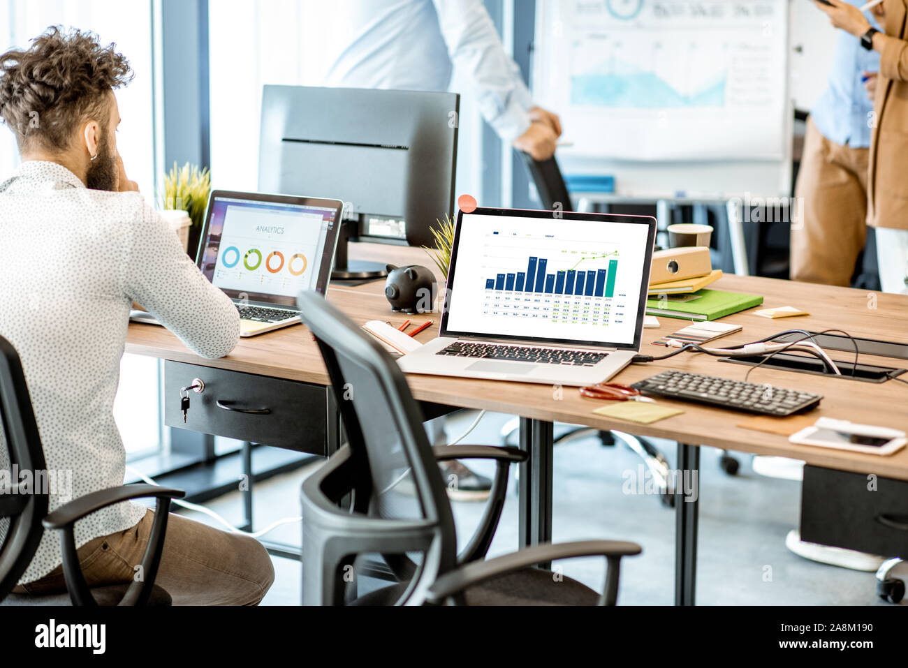 Uomo seduto sul posto di lavoro con computer, pensiero su alcuni finanziario tabelle e grafici in ufficio con altri colleghi Foto Stock