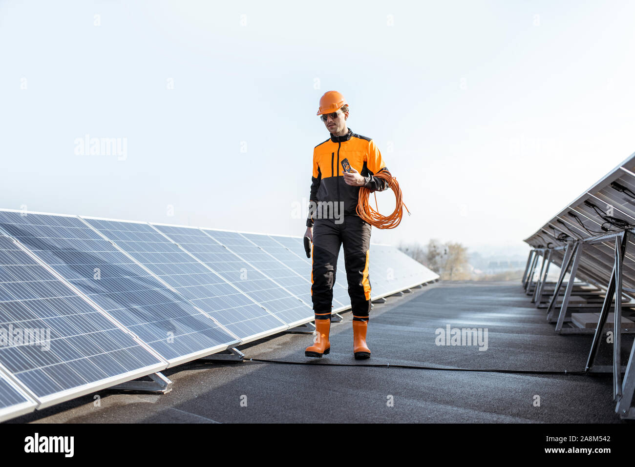 Ben attrezzato lavoratore in arancione di protezione abbigliamento passeggiate ed esaminando i pannelli solari su un tetto fotovoltaico impianto. Concetto di manutenzione e installazione di stazioni solari Foto Stock