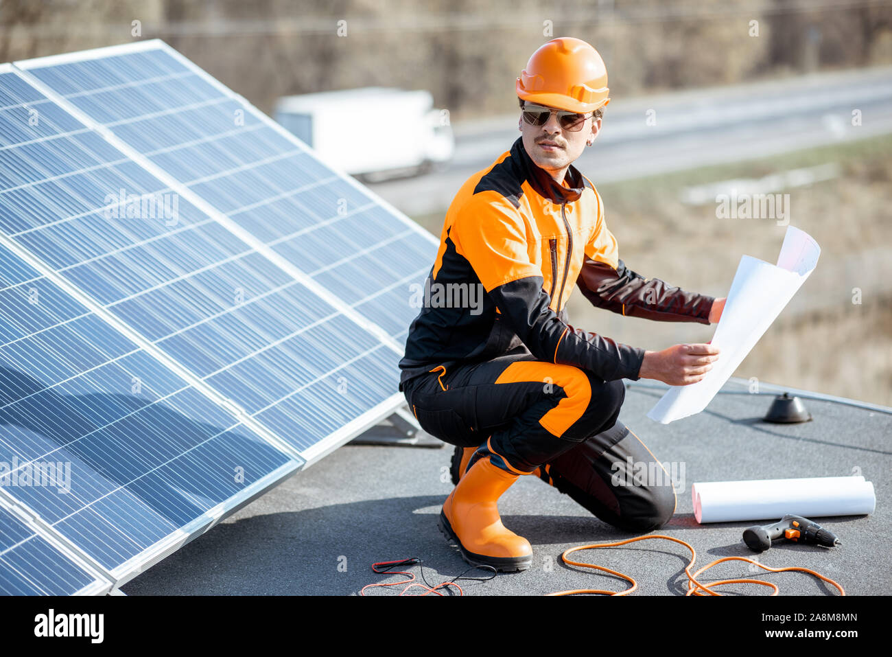 Elettricista in indumenti da lavoro protettiva di installare pannelli solari, seduto su un tetto con blueprint e strumenti di lavoro Foto Stock