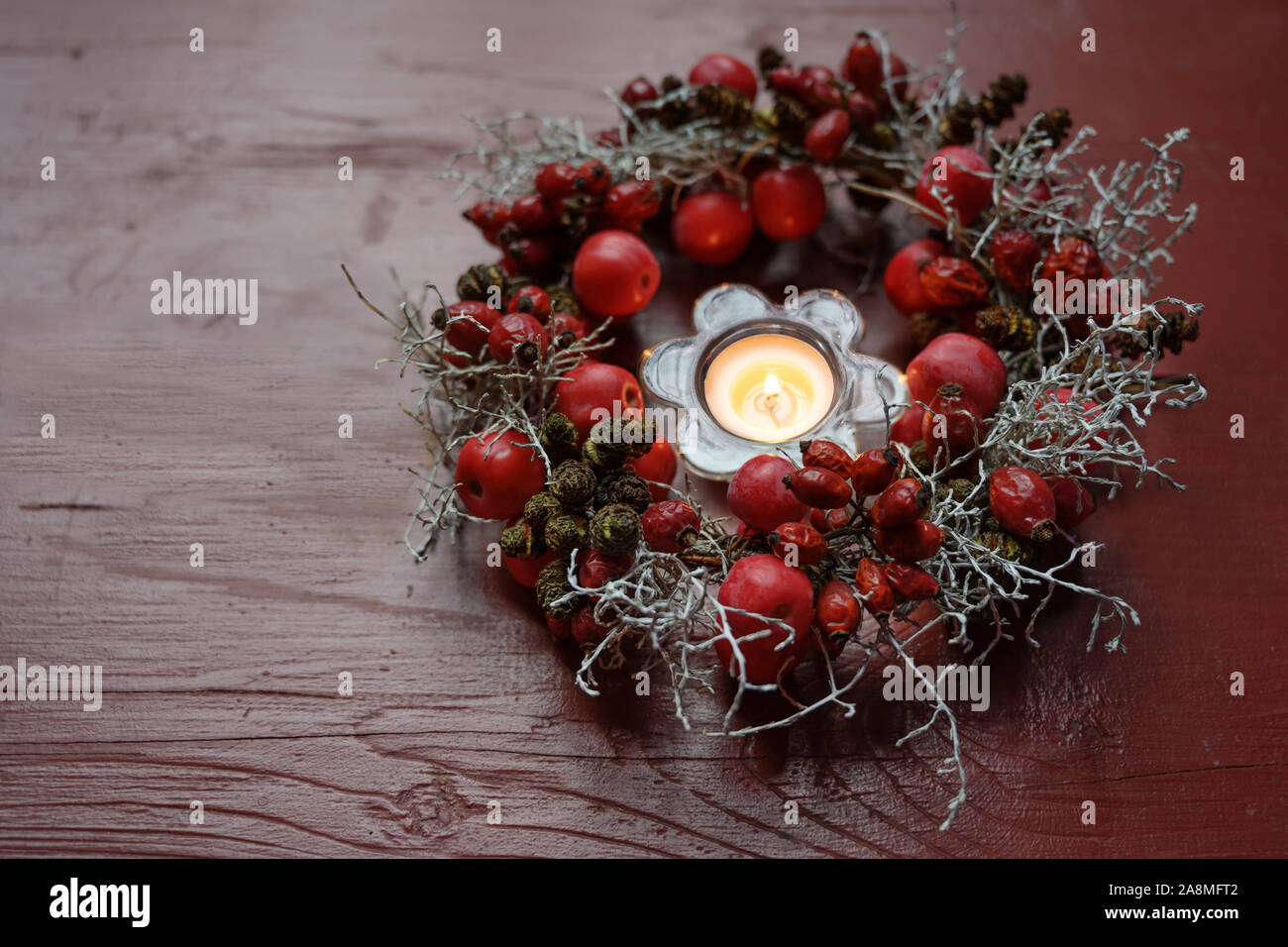 Ghirlanda di naturale con rosa canina e piccole mele intorno ad una candela che brucia in un rosso tavolo in legno, decorazioni stagionali in autunno, Avvento e Natale Foto Stock