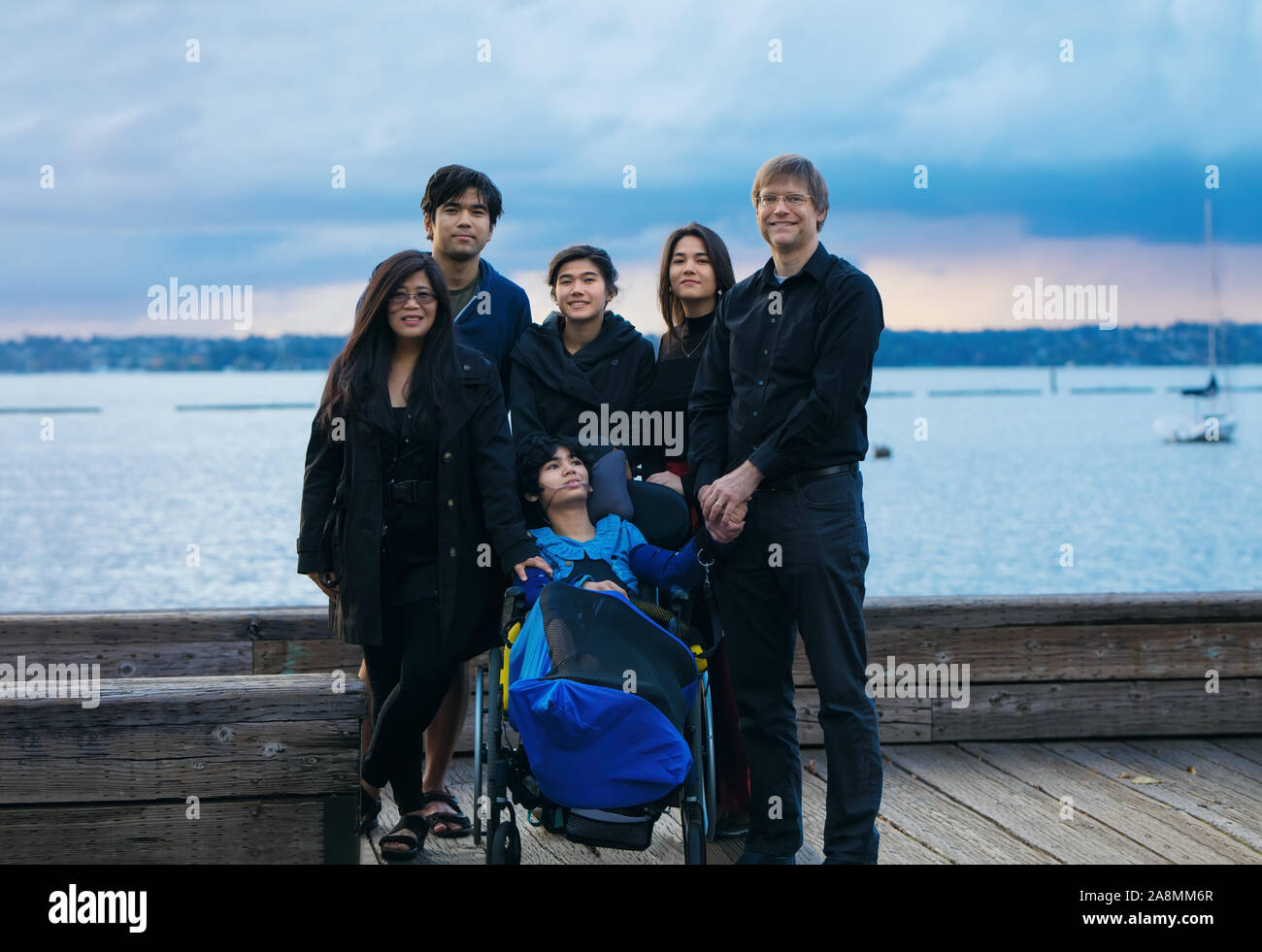 Razza mista famiglia con disabili little boy in sedia a rotelle all'aperto sul molo in legno dal lago sulla sera Nuvoloso al tramonto a inizio autunno Foto Stock