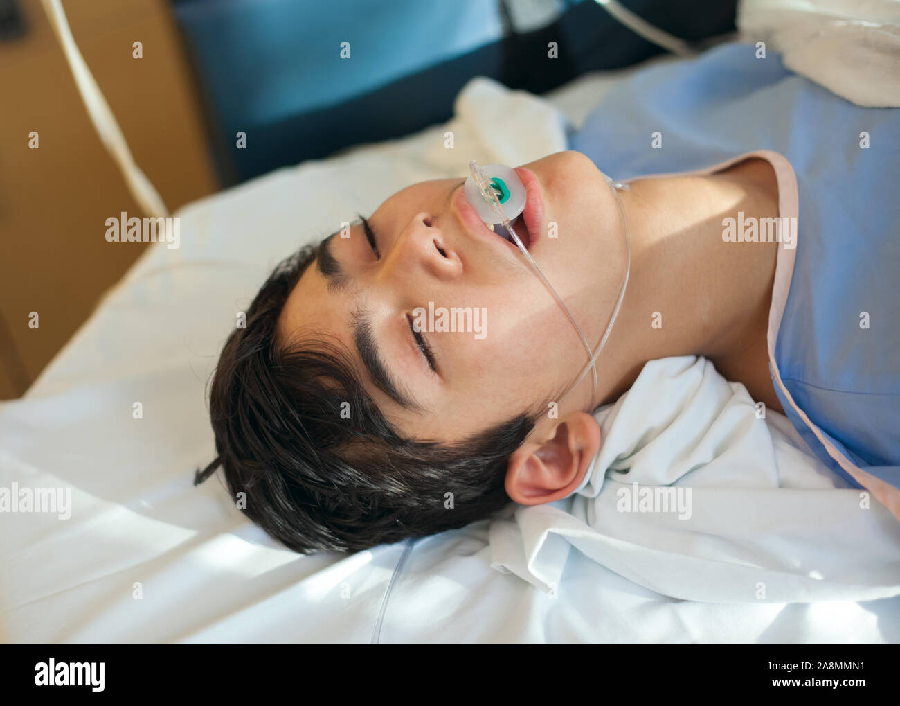 I giovani di tredici anni biracial disabili boy giacente in stato di incoscienza in ospedale gurney letto in camera di recupero indossando blue abito ospedale, tubo di respirazione dow Foto Stock