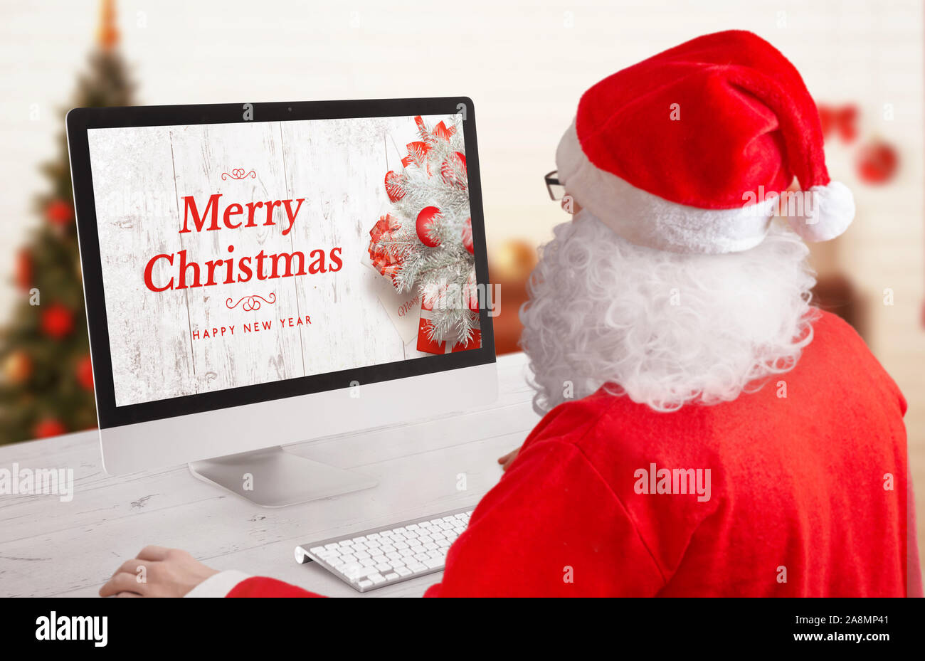 Santa inviare Natale e Anno Nuovo biglietto di auguri per posta dalla sua stanza di lavoro. Scena con un moderno computer di visualizzazione e di albero di Natale in background. Foto Stock