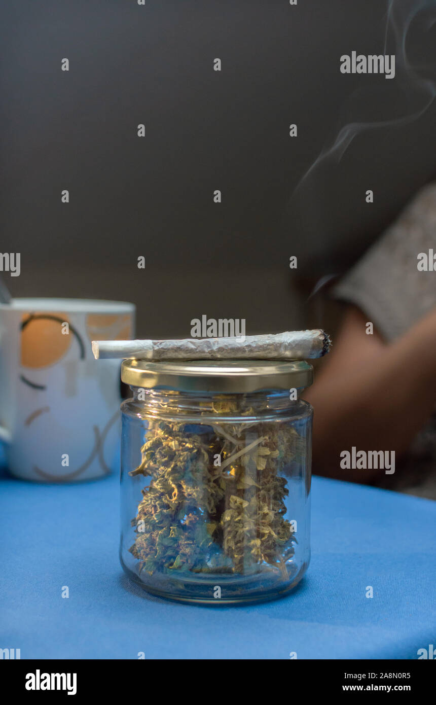 Fumare un giunto di marijuana con una tazza di caffè a casa. Riutilizzare il vasetto di vetro pieno di boccioli di cannabis con un erbaccia sigaretta, vicino. Copia superiore dello spazio. Foto Stock