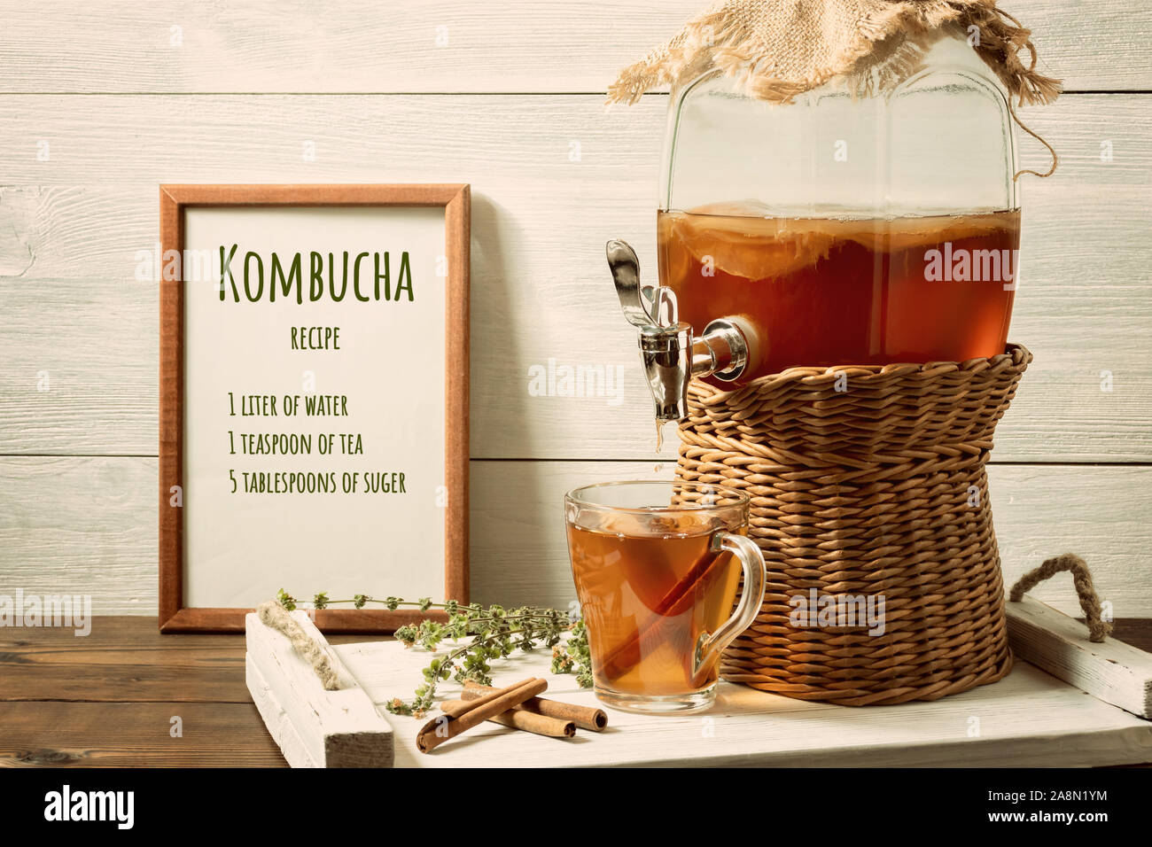 Freschi Fatti in casa il Kombucha tè fermentato da bere in un vasetto con rubinetto e in un bicchiere su un vassoio di bianco, la ricetta per la cottura nel telaio, sullo sfondo di legno. Foto Stock