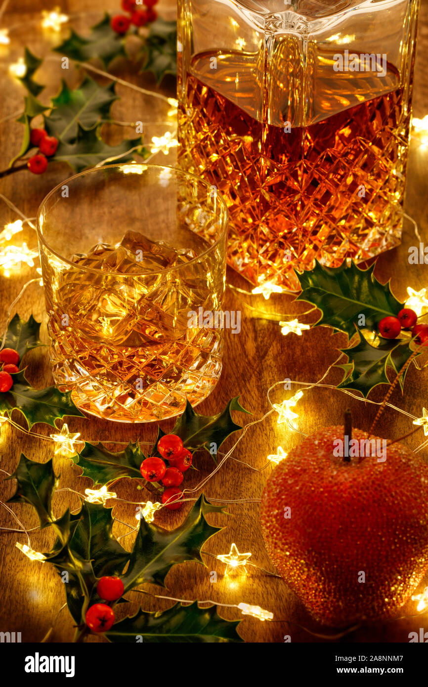 Crystal decanter di whisky e un bicchiere di whisky con ghiaccio, le luci di Natale e holly bacche, su di un tavolo di legno Foto Stock