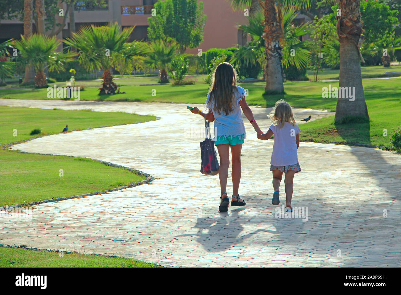 Piccoli turisti andare su strada in località tenendo la mano. Due piccole sorelle di andare sul percorso tra alberi di palma. Le giovani ragazze godendo le vacanze estive Foto Stock